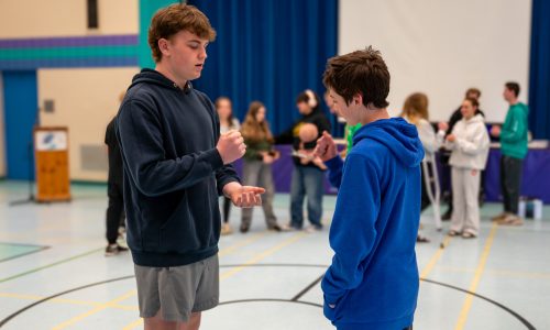 Two male students play a game of Rock, Paper, Scissors while standing in a school gymnasium.
