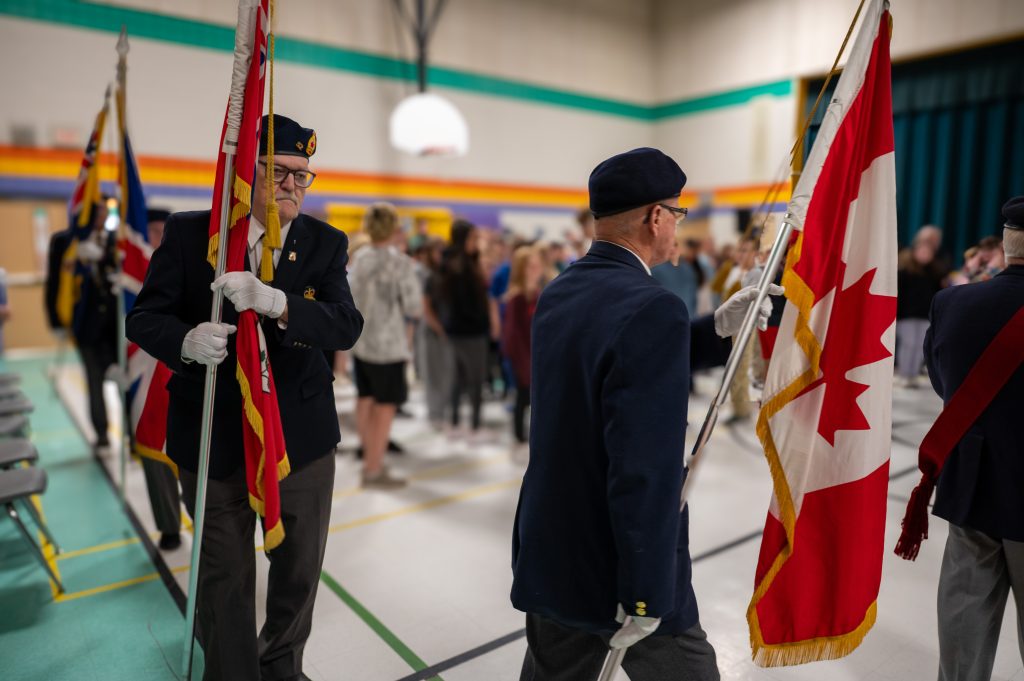 The Royal Canadian Legion marches in a procession carrying the colours while in a school gymnasium.