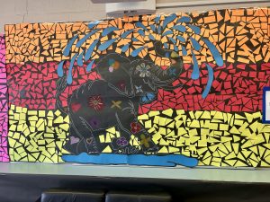 A mosaic display on a wall of an elephant spraying water out of its trunk created using smaller colourful pieces of paper.