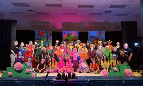A large group of students wearing colourful costumes gather on stage for a musical.