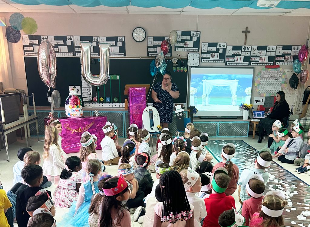 A classroom of students sits on the floor for the make believe wedding of the letters "Q" and "U".