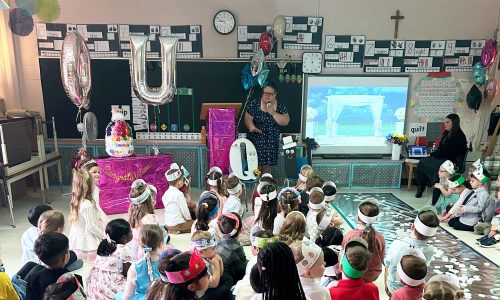 A classroom of students sits on the floor for the make believe wedding of the letters "Q" and "U".