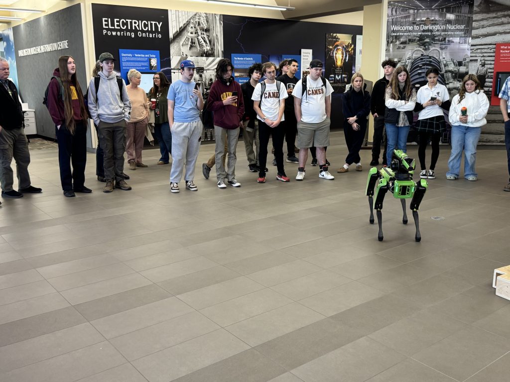 Students gather around in a circle while a green robot does tricks at a nuclear power generating station.