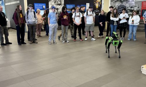 Students gather around in a circle while a green robot does tricks at a nuclear power generating station.