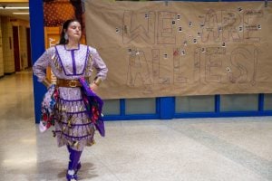 An Indigenous woman performs a Traditional Pow Wow Jingle Dress Dance in a high school foyer.