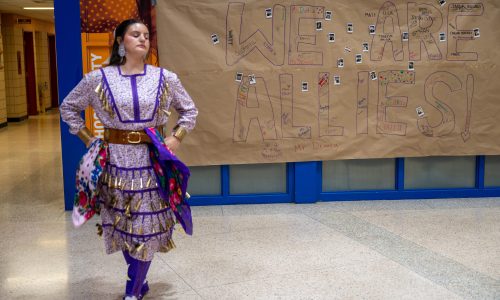 An Indigenous woman performs a Traditional Pow Wow Jingle Dress Dance in a high school foyer.