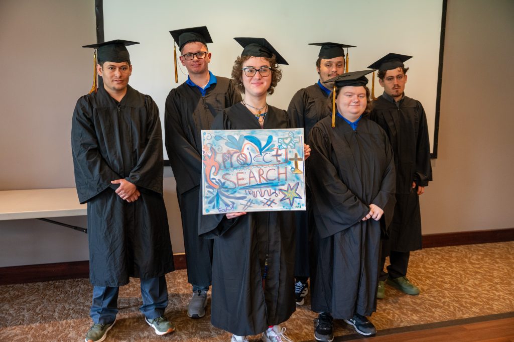 Six students, dressed in black graduation gowns, hold up a sign that reads "Project Search".