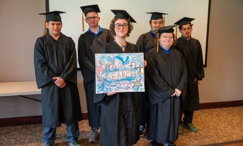 Six students, dressed in black graduation gowns, hold up a sign that reads "Project Search".