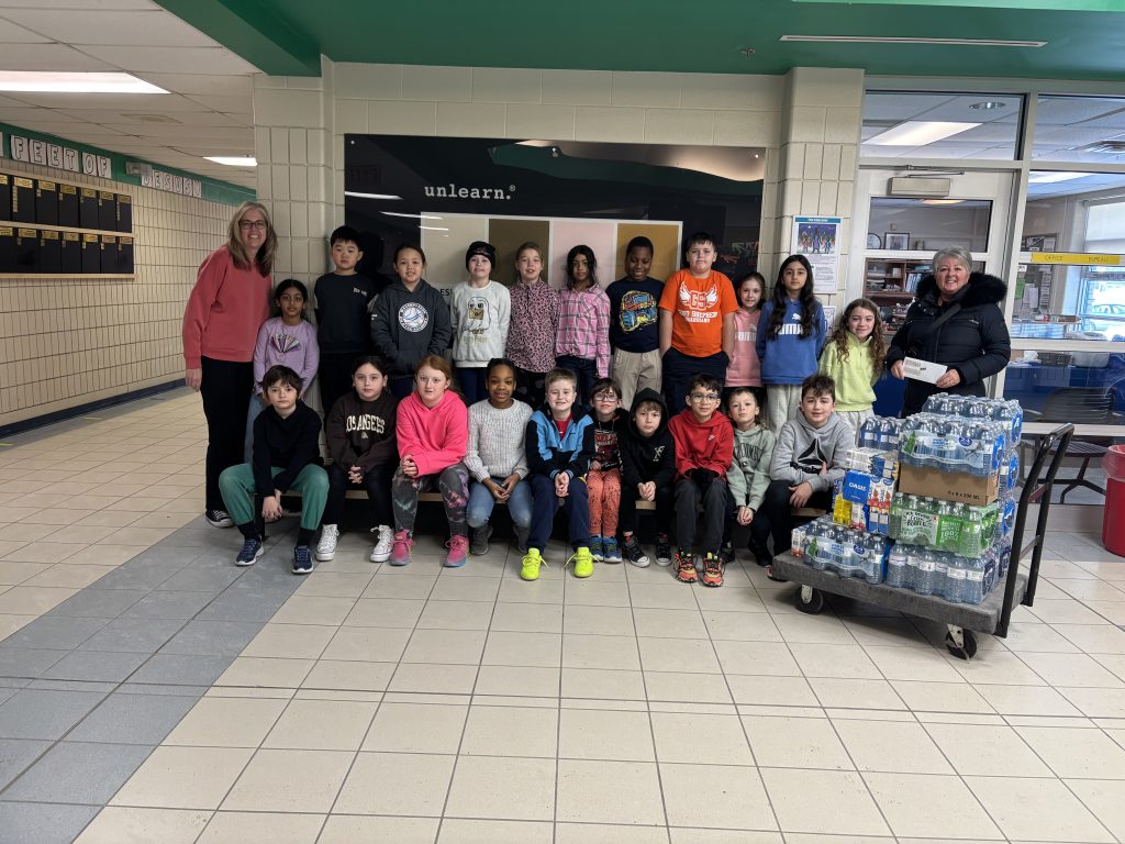 Twenty one students stand with two adults and a trolly with cases of water on it.