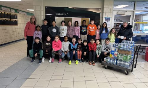 Twenty one students stand with two adults and a trolly with cases of water on it.
