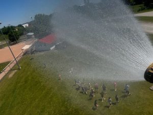 A firefighter stands in the bucket at the top of the ladder on a pump truck spraying water onto children below as they keep cool at a baseball game.
