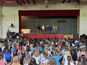 Six students dressed up as animals perform a song and dance on a stage in a school gymnasium in front of a large crowd of students.