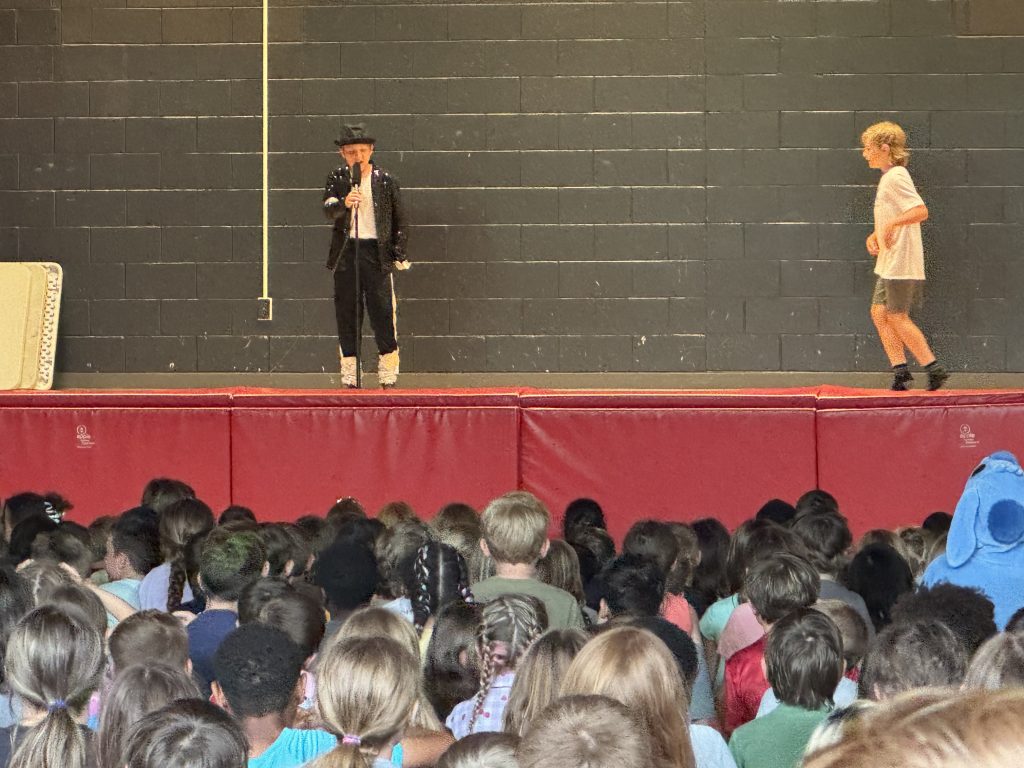 Two students perform a tribute to Michael Jackson on a stage in a school gymnasium in front of a crowd of students.