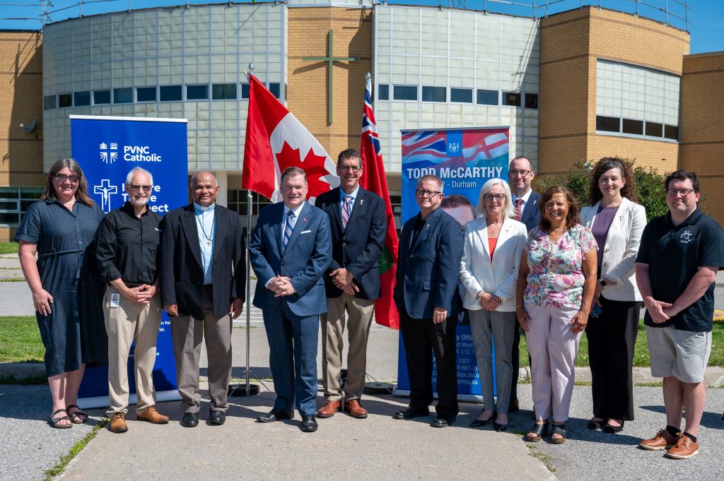 Eleven adults stand in front of two large pull-up banners, a Canadian and an Ontario flag in a school parking lot for a press conference.