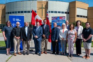 Eleven adults stand in front of two large pull-up banners, a Canadian and an Ontario flag in a school parking lot for a press conference.