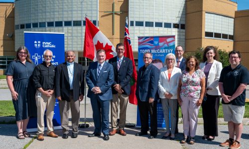 Eleven adults stand in front of two large pull-up banners, a Canadian and an Ontario flag in a school parking lot for a press conference.